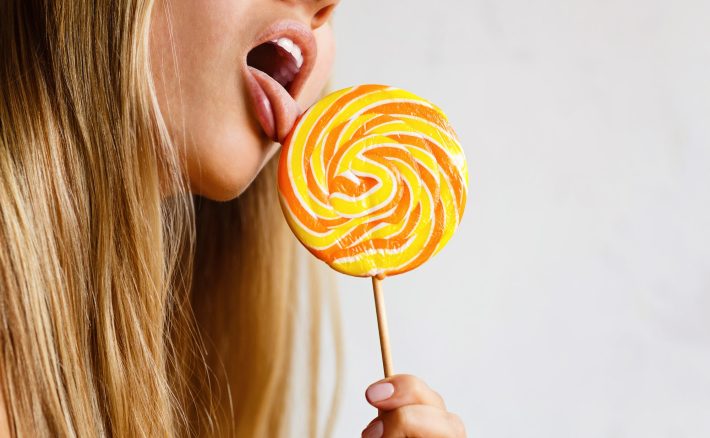 Young Caucasian woman with long blonde hair licking bright sweet candy on gray background